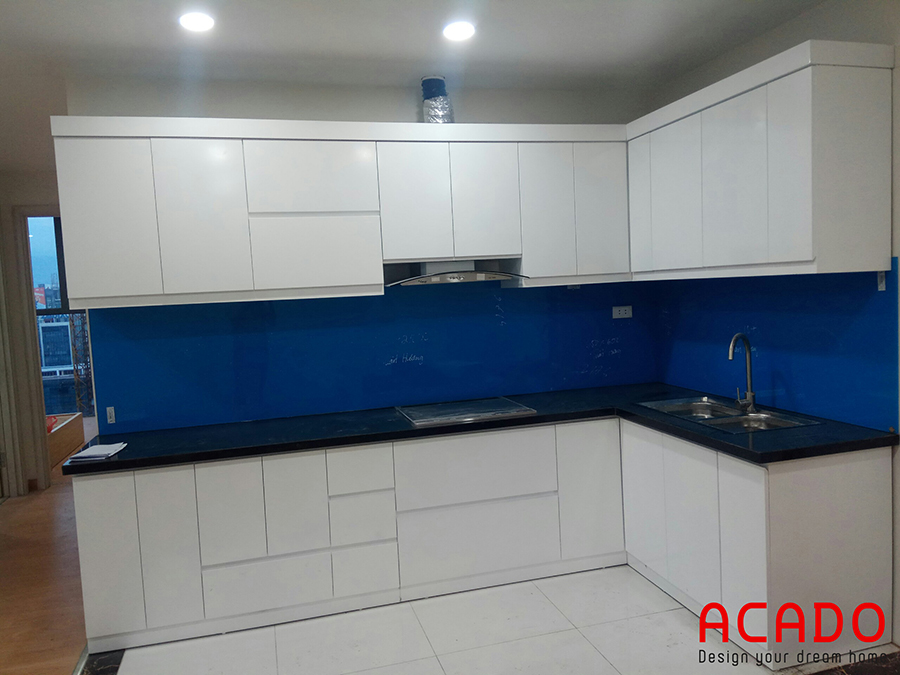Tủ bếp Melamine hình chữ L màu trắng với điểm nhấn là kính ốp màu xanh cho không gian bếp thoải mái và tiện nghi